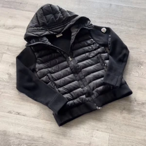 Svart pufferjacka från Moncler - Säljer en svart pufferjacka från Moncler med huva och ribbstickade ärmar. Jackan har quiltad framsida, tvåvägsdragkedja och Moncler-logga på axeln. Materialmix med polyester och stickade detaljer ger en cool och sportig vibe.