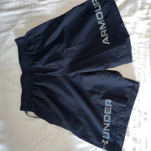 Svarta träningsshorts från Under Armour - Svarta träningsshorts från Under Armour med elastisk midja och stort logotryck på ena benet. Modellen är loose fit och tillverkad i lätt polyester som andas, perfekt för gymmet eller löprundan.