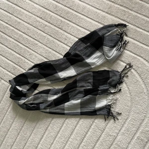 Svartvit rutig halsduk med fransar - Snygg halsduk i svart, vitt och grått med rutigt mönster och tunna fransar i ändarna. Lätt och luftig, perfekt att slänga över axlarna för en cool look. Passar till de flesta outfits och ger en stilren touch.