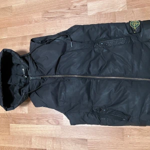 Svart pufferjacka från Stone Island - Svart pufferjacka från Stone Island med huva och dragkedja framtill. Jackan har den klassiska Stone Island-loggan på ärmen och praktiska fickor med knappdetaljer. Perfekt för dig som vill ha en clean och stilren look under kyliga dagar.