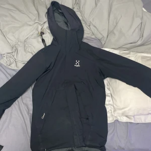 Svart vindjacka från Haglöfs herr S - Svart vindjacka från Haglöfs i herrmodell, storlek S. Jackan har huva med dragsko, meshfoder på insidan och dragkedja framtill. Tillverkad i slitstark polyester och perfekt för blåsiga dagar. Snygg minimalistisk design med diskret logga på bröstet.