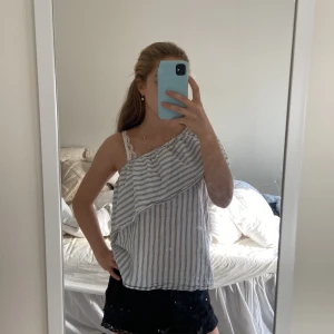Randig oneshoulder topp från H&M - Säljer en fräsch vit och blå randig oneshoulder topp från H&M i storlek 38. Toppen har en volangdetalj framtill och är tillverkad i ett lätt och luftigt linnematerial.