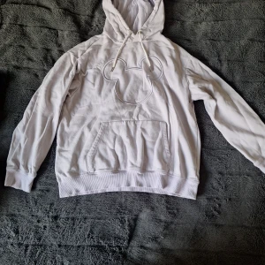 Lila  hoodie med Mickey Mouse-tryck - Lila hoodie från Disney med ett stort, broderat Mickey Mouse-ansikte på bröstet. Tröjan har huva med snörning, magficka och ribbade muddar vid ärmslut och nederkant. Perfekt för dig som gillar ikoniska motiv och avslappnad stil.