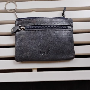 Svart skinnplånbok från Feel - Minimalistisk svart plånbok i äkta skinn från Feel med dragkedja och extra fack framtill. Praktisk nyckelring på insidan gör det enkelt att hålla koll på nycklar och småprylar. Perfekt storlek för fickan eller väskan.