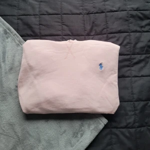 Ljusrosa hoodie från Ralph Lauren - Snygg rosa hoodie från Ralph Lauren med klassisk blå broderad logga på bröstet. Tröjan har huva med snörning. Perfekt för chill dagar och enkel att matcha med jeans eller joggers.