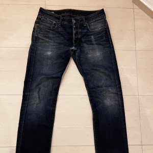 Mörkblå jeans från G-Star RAW - Snygga mörkblå jeans från G-Star RAW med slitna detaljer och kontrastsömmar. Modellen är mer slim fit men lite straight leg🙌Jeansen har flera lagningar som berättar en livshistoria🕰️. Jeansen är tillverkade i robust denim men med åren blivit lite svagare✌️Storlek W33 L32