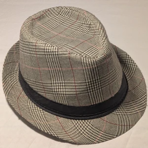 Rutig hatt - Snygg fedora hatt i klassiskt rutigt mönster med inslag av beige, svart och röda linjer. Hatten har ett svart band runtom och en stilren form som ger en cool vibe. Perfekt för dig som vill sticka ut med en unik accessoar. Materialet känns som bomull eller syntet.