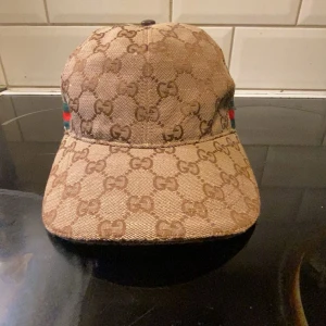 Gucci keps med GG-mönster och ränder - Säljer en snygg beige keps från Gucci med klassiskt GG-mönster över hela kepsen. På sidorna finns gröna och röda ränder som ger extra lyxig känsla. Justerbar rem baktill och böjd skärm. Perfekt för dig som vill sticka ut med designerstil.