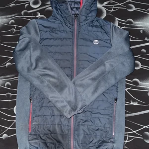Mörkblå pufferjacka från Jack - Snygg mörkblå pufferjacka med huva från Jack. Jackan har quiltad framsida och rygg, röda detaljer på dragkedjorna och två sidofickor med dragkedja. Ärmarna är släta och materialet känns mjukt och sportigt. Perfekt för kyliga dagar. Storlek har en i 164 och en i 158
