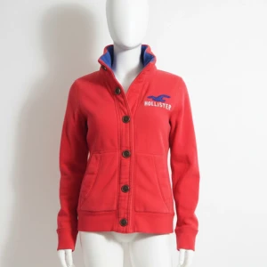 Röd kofta från Hollister med knappar - Röd kofta från Hollister med blå logga och text på bröstet. Tröjan har hög krage med blå insida, stora knappar framtill och två fickor. Perfekt för dig som gillar en sportig och avslappnad stil.