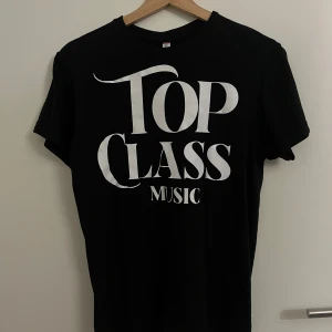 Svart Top Class Music t-shirt - Svart t-shirt med stor vit text 'Top Class Music' på framsidan. Klassisk rund hals och korta ärmar. Passformen är normal och materialet känns som mjuk bomull, perfekt för dig som gillar musik och vill ha en enkel, stilren look.