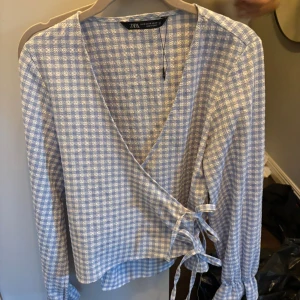 Blåvit rutig omlottblus från Zara - Säljer denna söta blåvit rutiga blusen från Zara med knyt i sidan och långa ärmar. Blusen har v-ringad hals och är använd en gång innan