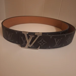 Louis Vuitton svart skinnbälte - Svart skinnbälte från Louis Vuitton med klassiskt monogrammönster och stor LV-spänne i silver. Bältet har en stilren och exklusiv look med mörka toner och detaljerad präglad insida. Perfekt för dig som vill ha en lyxig accessoar med tydlig designerstil.