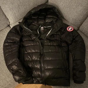 Svart dunjacka Canada Goose Crofton -  Storlek S. Svart Crofton Hoody dunjacka från Canada Goose med huva, ikonisk patch på ärmen och quiltad design. Jackan har dragkedja framtill, ribbade muddar och lätt glansigt material. Perfekt för kalla dagar och har snygg streetstil. Har ett trådsmalt hål på vänster arm på bilden, inget bekymer eller något man lägger märke till enligt mig, i övrigt bra skick. 