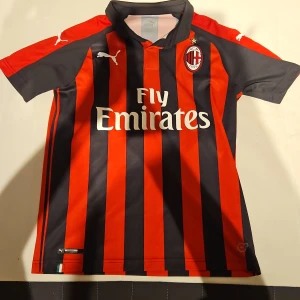 AC Milan fotbollströja Puma röd/svart - AC Milan matchtröja från Puma med klassiska röda och svarta vertikala ränder, klubbmärke på bröstet och Fly Emirates-tryck. Kortärmad modell i lätt polyester, med italiensk flaggdetalj vid nederkant och vit Puma-logga på axlarna.
