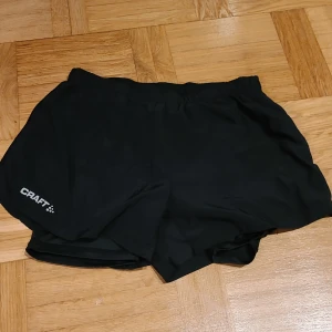 Svarta träningsshorts från Craft - Svarta träningsshorts från Craft i storlek S. De har elastisk midja, liten dragkedjeficka bak och reflekterande detaljer på sidorna. Perfekta för löpning eller gym, tillverkade i lätt syntetmaterial som andas.