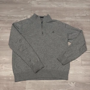 Grå half zip tröja från GANT - Snygg grå tröja från GANT med halv dragkedja och broderad logga på bröstet. Tröjan är ribbstickad vid krage, ärmslut och nederkant och är tillverkad i mjuk bomull. Perfekt för en stilren och avslappnad look.