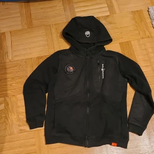 Svart hoodie från Sail Racing med huva - Svart hoodie från Sail Racing med dragkedja framtill, broderad logga på bröstet och en bröstficka med dragkedja. Tröjan har huva, långa ärmar och förstärkta partier på armbågarna. Perfekt för dig som gillar sportig och stilren look.