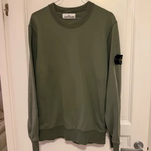 Olivgrön sweatshirt från Stone Island - Säljer en olivgrön sweatshirt från Stone Island med klassisk rund hals och ikonisk logga på vänster ärm. Tröjan har en clean look, ribbade muddar och är perfekt för dig som gillar streetwear. Materialet är mjuk bomull och passformen är normal.