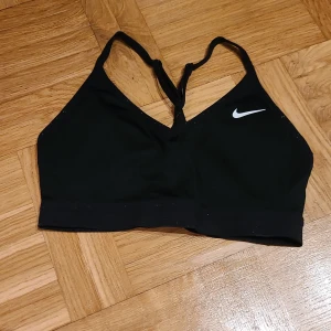 Svart sportig linnetopp från Nike - Svart linnetopp från Nike med smala axelband som korsas i ryggen. Toppen har en enkel och sportig design med vit Nike-logga framtill. Perfekt för träning och tillverkad i stretchigt syntetmaterial för maximal komfort.