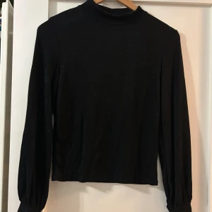 Svart glittrig polotopp - Snygg svart polotopp med diskret glittrigt tyg och långa ärmar. Toppens höga krage ger en stilren look och materialet är mjukt och följsamt. Perfekt för dig som vill ha något enkelt men ändå med lite extra shine.