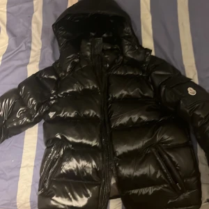 Svart Moncler pufferjacka med huva - Säljer en svart, glansig pufferjacka från Moncler med huva och dragkedja. Jackan har klassisk Moncler-logga på ärmen och är vadderad för extra värme. Perfekt för kalla dagar och har en riktigt clean och modern look.NFC tag är med size 3