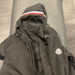 Svart vindjacka från Moncler - Svart vindjacka från Moncler med huva och dragkedja. Jackan har Moncler-logga på ärmen och en snygg rand i rött, vitt och blått på huvan. Tillverkad i lätt och glansig polyester, perfekt för dig som gillar stilren och sportig look. Storlek S, mer info skriv , Sista pris 1200 tyvärr . Skick 8,5/10