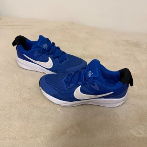 blå sneakers - Snygga Nike Star Runner sneakers i klarblå färg med vita detaljer och svart häl. Skorna har en sportig design med snörning, rund tå och platt sula. Ovandelen är tillverkad i syntetmaterial med mesh för extra ventilation. Perfekta för dig som gillar en aktiv stil. Skorna kommer skickas ut rena.