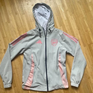 Arsenal x Adidas vindjacka ljusgrå/rosa - Snygg ljusgrå vindjacka från Adidas i samarbete med Arsenal. Jackan har rosa detaljer, huva med snörning, dragkedja framtill och elastiska muddar. Perfekt för dig som vill ha sportig stil och visa vilket lag du hejar på.