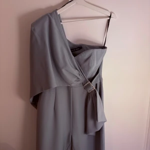 Ljusgrön oneshoulder klänning Lavish Alice - Supersnygg ljusgrå långklänning från Lavish Alice med oneshoulder-design och elegant drapering över axeln. Klänningen har en glittrig dekorerad spänndetalj i midjan som ger extra lyxig känsla. Perfekt för dig som vill sticka ut med stilren look.