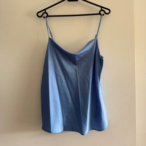 Ljusblå linnetopp från Samsara - Supersnygg ljusblå linnetopp från Samsara med tunna justerbara axelband och lätt glansigt tyg. Toppen har en avslappnad passform och en mjukt draperad halsringning som ger en stilren look. Perfekt för varma dagar eller att styla lager på lager.