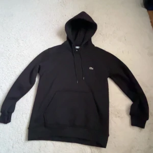 Svart hoodie från Lacoste med huva - Svart hoodie från Lacoste i storlek M med klassisk krokodillogga på bröstet. Tröjan har huva med snörning, stor magficka och långa ärmar. Materialet är mjuk bomull med stretch för extra komfort. Perfekt för chill dagar och streetwear. Helt ny hoodie, inte använd en ända gång. 