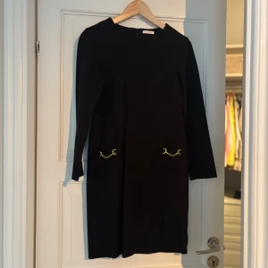Svart klänning med gulddetaljer Ida Sjöstedt - Svart klänning med lång ärm och rund hals. Två fickor framtill pryds av snygga guldfärgade kedjedetaljer som ger en cool touch. Klänningen har en rak och enkel siluett som känns både stilren och modern.