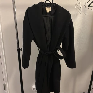 Svart kappa med bälte från hm - Stilren svart kappa från hm med bälte i midjan och bred krage. Den är lite damig men de går nog att ta bort