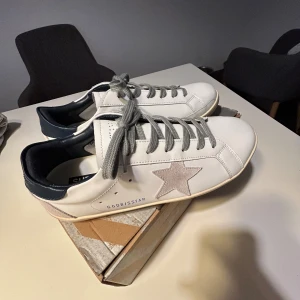 Golden Goose Superstar vita sneakers - Golden Goose Superstar sneakers i vitt skinn med grå snörning och en beige stjärndetalj på sidan. Mörkblå häl och platt sula ger en clean och cool look. Perfekt för dig som gillar stilrena och trendiga sneakers med unika detaljer.