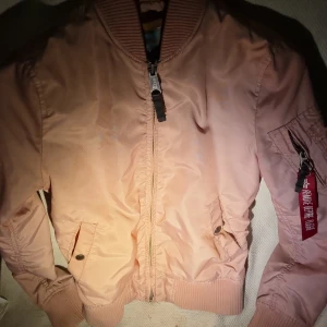 Rosa bomberjacka Alpha Industries XS - Snygg rosa bomberjacka från Alpha Industries i storlek XS. Jackan har klassisk ribbad krage, muddar och dragkedja framtill samt fickor med knapp. På ärmen finns en cool ficka med röd tag. Perfekt för dig som vill ha en streetig look.