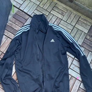 Svart Adidas zip up XL - Svart vindjacka från Adidas i storlek XL med klassiska vita ränder längs ärmarna och Adidas-logga på bröstet. Jackan har dragkedja framtill och är långärmad. Perfekt för sportiga dagar eller chill häng med kompisarna.