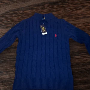 Blå stickad tröja från Polo Ralph Lauren - Kabelstickad blå tröja från Polo Ralph Lauren med halv dragkedja i halsen och klassisk röd logga broderad på bröstet. Tröjan har lång ärm och ribbade muddar för en snygg passform. Perfekt för lager-på-lager under kyliga dagar.