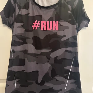 Camo t-shirt med #RUN tryck - Cool t-shirt med grå och svart camouflage-mönster och ett stort rosa #RUN-tryck på bröstet. Tillverkad i stretchig polyester och elastan, perfekt för träning. Kortärmad och med sportig passform som sitter snyggt på kroppen.