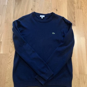 Mörkblå tröja från Lacoste - Säljer en klassisk mörkblå tröja från Lacoste med rund hals och den ikoniska gröna krokodilloggan broderad på bröstet. Tröjan har lång ärm och är tillverkad i mjuk bomull, perfekt för en stilren och enkel look. Tröjan är i storlek XS och passar för 160-170. 
