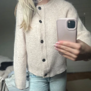 Beige stickad kofta med svarta knappar - Mysig beige stickad kofta med svarta knappar framtill. Koftan har en rak passform och långa ärmar, perfekt för lager-på-lager. Den är enkel men ändå stilren och passar till jeans eller kjol.