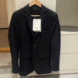 Marinblå kavaj Selected Homme 42 - Snygg marinblå kavaj från Selected Homme. Kavajen har två knappar framtill, klassiska fickor med lock och en bröstficka. Slim passform och stilren design, perfekt för dig som vill ha en modern look. Aldrig använd, prislappen kvar. 