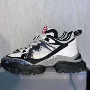Chunky sneakers från Moncler, vit/svart - Säljer ett par leave no trace sneakers från Moncler i vitt, svart och silver. Skorna har grov sula, snörning och coola detaljer i mesh, skinn och syntet. Röd, vit och blå Moncler-flärp på plösen ger extra stilpoäng. Perfekta för dig som gillar statement-sneakers. Låda och dustbags medföljer.