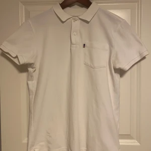 Lexington piké - Klassisk vit pikétröja från Lexington i storlek S. Tröjan har korta ärmar, krage, knäppning med två knappar och en bröstficka. Tillverkad i mjuk bomull som känns skön mot huden. Perfekt för en clean och stilren look.