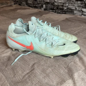 Nike fotbollsskor ljusblå US 11 - Säljer ett par Nike fotbollsskor i ljusblått med snörning och orange Swoosh på sidan. Skorna har platt sula med dobbar för bra grepp på planen och är tillverkade i syntetmaterial. Perfekta för dig som vill sticka ut på fotbollsplanen.