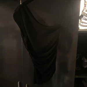 Svart oneshoulder kortklänning - Snygg svart kortklänning med oneshoulder-design och draperad effekt längs sidan. Perfekt för dig som vill sticka ut med en stilren och trendig look. Klänningen har en tight passform och är tillverkad i ett mjukt, stretchigt material.