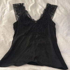 Svart linnetopp med spetsdetaljer - Supersnygg svart linnetopp med breda axelband och v-ringning, dekorerad med blommig spets framtill och längs axelpartiet. Materialet är mjukt och glansigt, vilket ger en lyxig känsla. Perfekt för dig som gillar stilrena och feminina detaljer. Den är figursydd och fin från MSCH copenhagen.