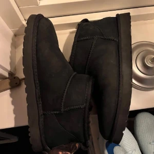 Svarta UGG - Säljer ett par klassiska svarta UGG boots med rund tå och platt sula. Skorna är tillverkade i mjuk mocka och har en robust yttersula som passar perfekt för kyliga dagar. UGG-loggan syns tydligt baktill. Grymt sköna och stilrena boots för höst och vinter.