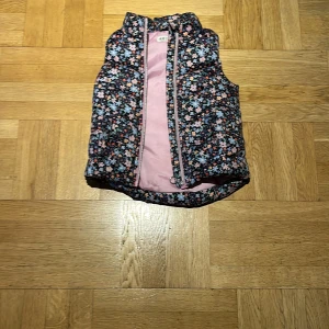 Blommig dunväst från H&M - Söt dunväst från H&M med blommigt mönster i ljusblått, rosa, orange och beige på mörkblå botten. Insidan är ljusrosa och västen har dragkedja framtill. Perfekt för lager-på-lager och ger en färgglad touch till din outfit.