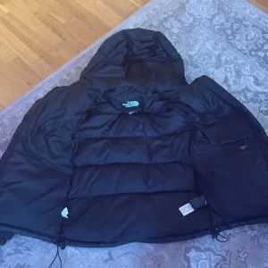 Svart dunjacka från The North Face XS - Säljer en svart dunjacka från The North Face i storlek XS. Jackan har huva med snörning, dragkedja framtill och stora fickor. Klassisk pufferstil med logga på bröstet och ryggen. Perfekt för kalla dagar och riktigt snygg streetstil.